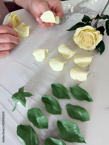 Handmade tea rose sculpted from air-dry clay lying on a table, with separate petals and green leaves nearby. Clay flower during crafting process. DIY art, handmade decoration, creative workshop