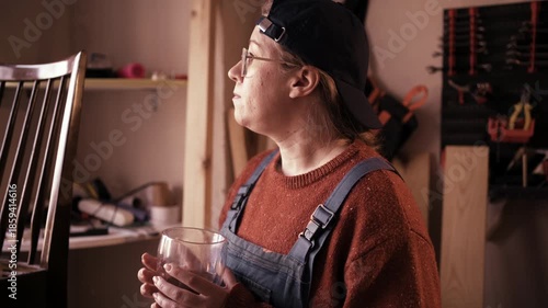 Young female craftsperson in overalls drinking coffee at workshop workplace