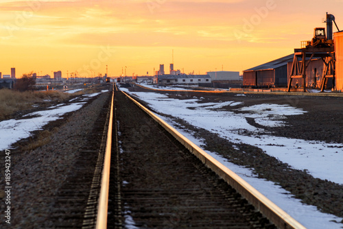 Railroad tracks run through an industrial area at sunset in Amarillo, Texas. Warehouses and industrial buildings line the rail corridor as warm light reflects off the rails. 