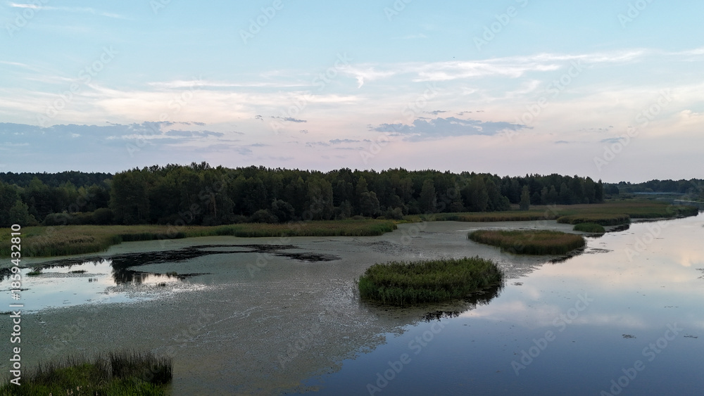 Fototapeta premium A serene view of a wide river surrounded by lush greenery under a pastel sky at dusk. Reflections on the water create a tranquil atmosphere, perfect for nature and tourism themes.