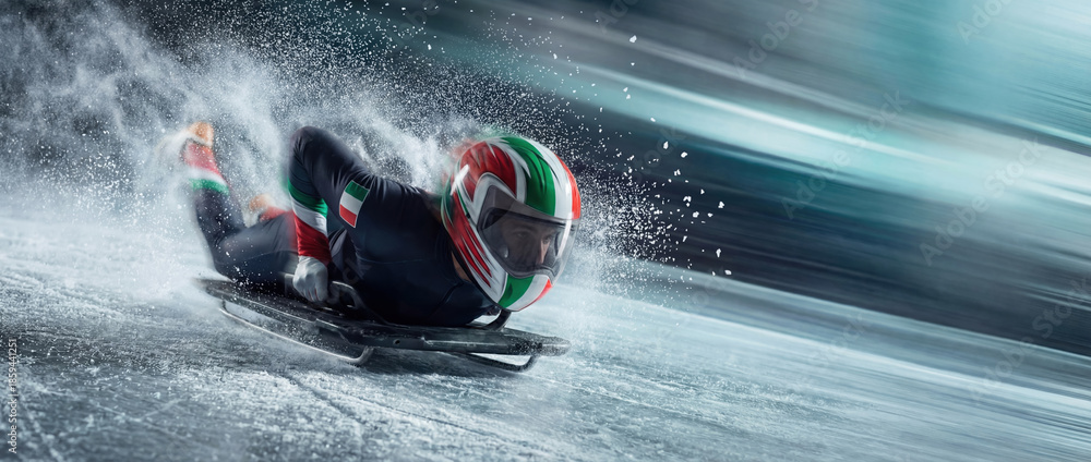 Fototapeta premium Skeleton athlete with an Italian helmet speeding down an ice track. Dynamic winter sport action shot with high-speed motion blur and flying ice spray for the 2026 games concept.