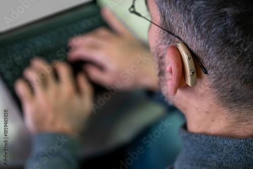 Man typing on a laptop keyboard, wearing a hearing aid. Illustrating concepts of accessibility and technology for people with hearing impairment