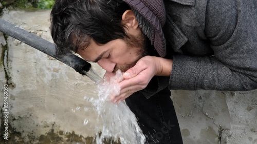 Young bearded man cupping hands to drink cool, pure water flowing from a natural spring pipe, quenching thirst outdoors in a serene, healthy rural environment