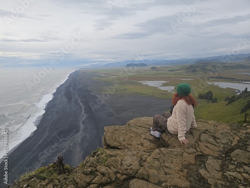 Woman with red hair on a cliff overlooking a vast black sand beach in Iceland. Cinematic volcanic landscape with endless coastline under a moody sky. Travel and solitude concept.