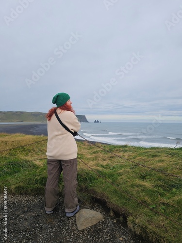 Woman with red hair looking at Reynisfjara black sand beach in Iceland. Stunning coastal landscape with sea stacks and moody sky. Travel and wanderlust concept.
