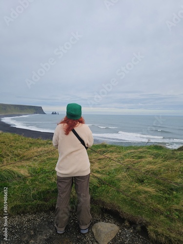 Woman with red hair looking at Reynisfjara black sand beach in Iceland. Stunning coastal landscape with sea stacks and moody sky. Travel and wanderlust concept.