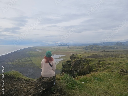 Woman with red hair sitting on a cliff in Iceland looking at a vast black sand coast and volcanic plains. Stunning Nordic landscape with mountains under a cloudy sky. Solitude and travel concept.