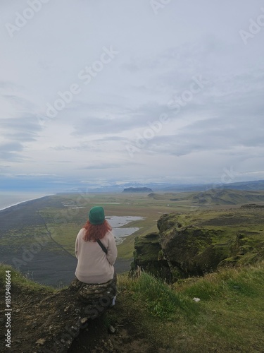 Woman with red hair looking at Reynisfjara black sand beach in Iceland. Stunning coastal landscape with sea stacks and moody sky. Travel and wanderlust concept.
