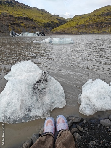 POV of traveler's feet by a glacial lagoon in Iceland with large floating icebergs and mossy mountains. Dramatic Nordic landscape and adventure travel concept.