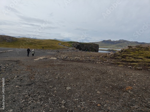 Tourists walking on a rocky path towards the Reynisfjara beach viewpoint in Iceland. Wide volcanic landscape with green hills and cloudy sky. Nordic travel and trekking concept.