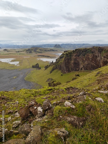 Vertical view from Dyrhólaey cliffs, Iceland. Mossy rocks in the foreground overlook a black sand beach, wetlands, and distant mountains.