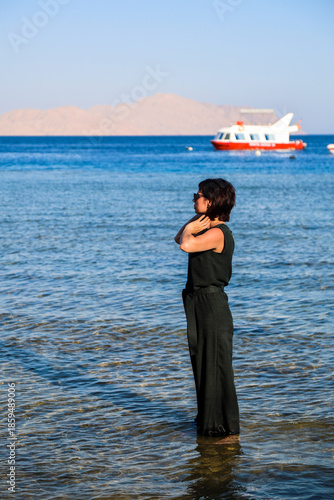 A grown woman in casual clothes stands knee deep in ocean water.