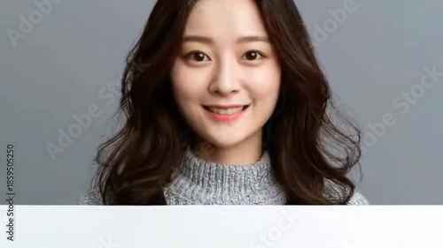 A smiling young adult female, with warm brown hair, confidently holds up a blank white sign against a clean, neutral gray background. Her cheerful and welcoming expression conveys positivity, engageme