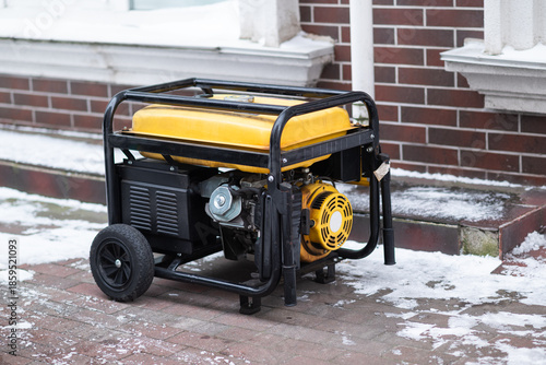 Yellow portable gasoline generator on wheels standing on a snowy sidewalk. Industrial backup power source for home or business during winter blackouts.