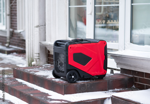 Red portable inverter generator standing on snowy stairs near a building. Backup power source for home during winter blackout or energy crisis.