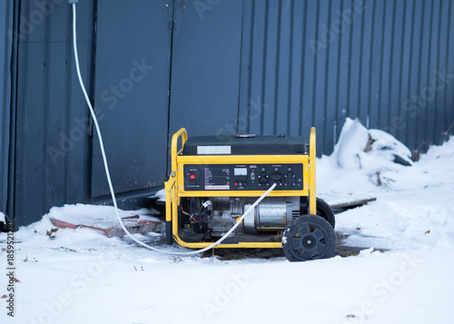 Portable yellow power generator standing outdoors in snow near an industrial building, winter emergency electricity supply concept.