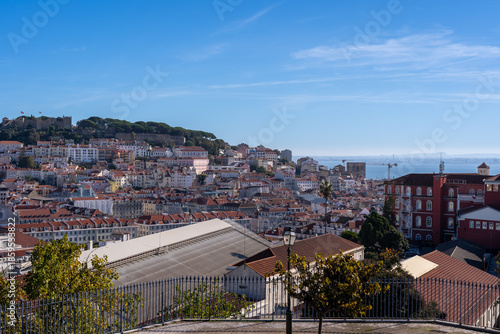High angle view of Lisbon, Portugal, featuring the historic Saint George Castle on the hill, traditional Mediterranean architecture with terracotta roofs under a clear blue sky.