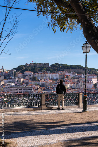 A male traveler standing at a viewpoint (Miradouro) looking over the historic city of Lisbon towards the castle. Lifestyle travel concept with a scenic urban background.