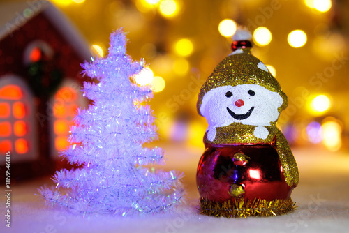 A snowman with a white Christmas tree and a small red house on a blurred background with yellow lights