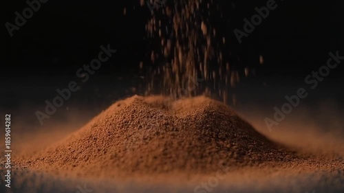 Close-up of a pile of brown powder with particles falling onto it against a dark background with warm lighting.