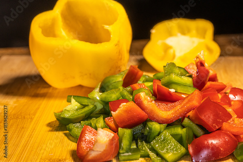 Fresh red, green, and yellow bell peppers are being chopped with a knife on a wooden cutting board. The scene shows healthy cooking, food preparation, and colorful vegetables in a kitchen setting.
