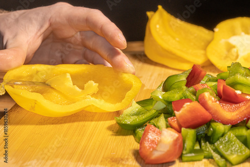 Fresh red, green, and yellow bell peppers are being chopped with a knife on a wooden cutting board. The scene shows healthy cooking, food preparation, and colorful vegetables in a kitchen setting.