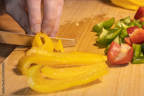 Fresh red, green, and yellow bell peppers are being chopped with a knife on a wooden cutting board. The scene shows healthy cooking, food preparation, and colorful vegetables in a kitchen setting.