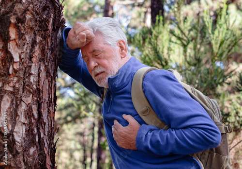 Tableau sur toile Senior man hiking in a forest pauses in distress leaning against a tree while holding his chest