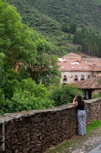A woman is taking a picture of a beautiful mountain view in village of Potes. The scene is peaceful and serene, with the woman standing on a stone wall overlooking the landscape