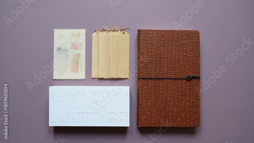 Artistic Flat Lay of Braille Slate, Textured Notebook, and Decorative Paper Elements on a Muted Purple Surface for Creative Projects and Visual Communication