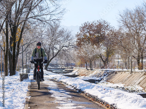 A cyclist rides along a city sidewalk in winter. An active winter lifestyle. An adult man on a red bicycle wearing a helmet