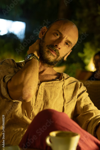 A portrait of a mature man sitting at a cozy outdoor cafe table during a warm summer evening, enjoying a cup of coffee. He looks relaxed and thoughtful in a modern urban lifestyle setting.