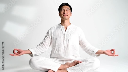 Asian man in white clothing meditating indoors in a lotus position against a clean white backdrop