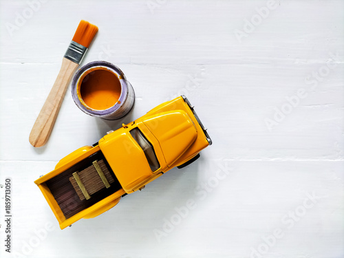 A small yellow toy car is placed on a white wooden surface beside an open jar of yellow paint and a paintbrush, highlighting the concept of repainting. 
