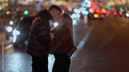 A young couple embraces on a street in Paris France at night The city lights are blurred in the background creating a romantic atmosphere The lovers share an intimate moment on a city street.
