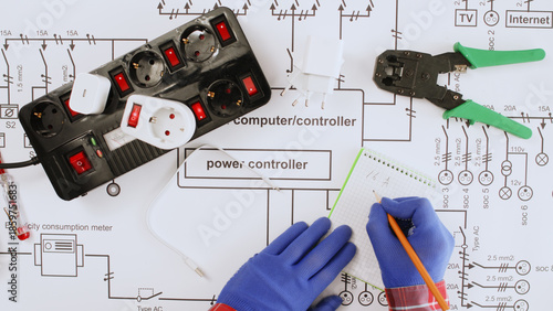 Obraz na plátně An electrical engineer writes down measurements on a large paper drawing of a house's electrical system, with a large uninterruptible power supply and network terminals lying nearby