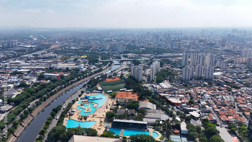 Obraz premium Vista aérea de cidade com parque aquático, avenidas, bairros densos, prédios e áreas verdes, mostrando lazer urbano, turismo, infraestrutura e expansão metropolitana.