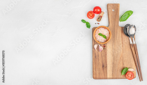 Minimal kitchen flat lay with wooden cutting board, tomatoes, basil, salt and utensils on white background with copy space