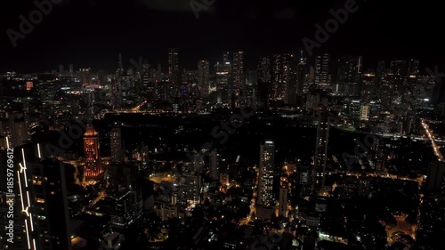 An aerial view of Mumbai's high-rise buildings and beautiful nightlights. The sky is cloudy, and there is medium traffic on the road. The cityscape is filled with modern illuminated buildings.