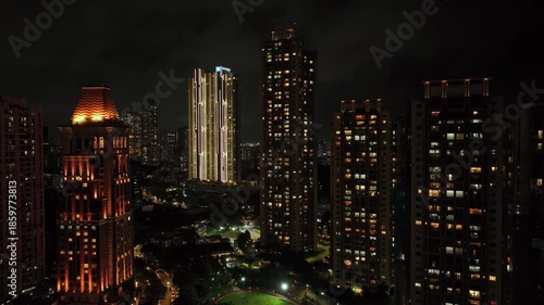An aerial view of Mumbai's high-rise buildings and beautiful nightlights. The sky is cloudy, and there is medium traffic on the road. The cityscape is filled with modern illuminated buildings.