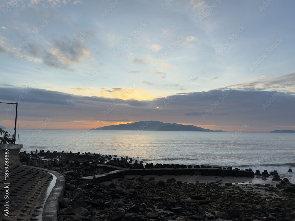 Fototapeta premium Sunrise Island Paradise: A distant island silhouette against a golden sunrise, with a rocky shore and calm ocean in the foreground under a blue sky with clouds.
