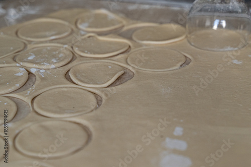 Perfectly cut dough circles dusted with flour on counter beside glass cutter, selective focus captures texture in cooking at home dumpling preparation process.