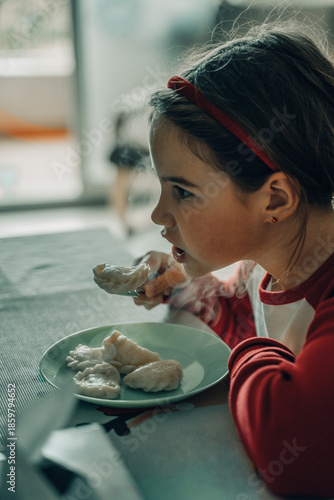 Cute girl with red headband savoring steamed dumplings from green plate at home table, selective focus on joyful expression captures cooking with grandma family moment