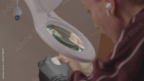 Focused artist using magnifying lamp and fine brush to paint delicate facial details on doll head in workshop