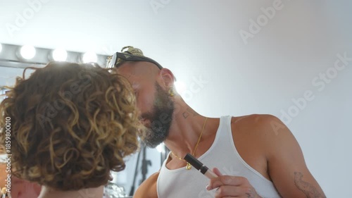 Focused bearded gay professional artist applying makeup on female model in illuminated dressing room before performance