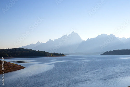 Sunbeams streaming down through hazy smoke haze of wildfire in front of mountains behind river lake under cloudless blue sky during blue hour of early morning