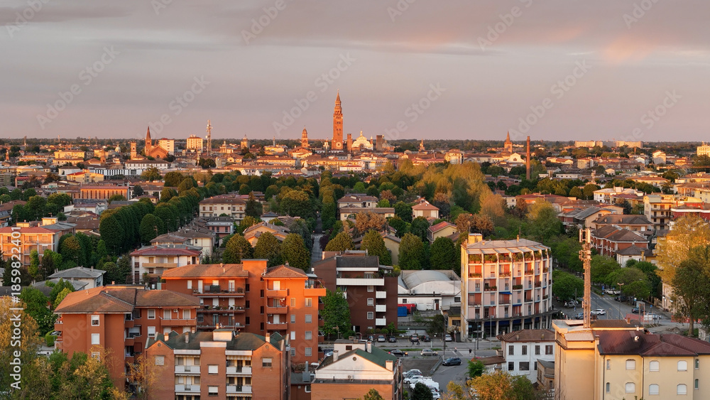 Fototapeta premium Aerial panorama revealing Cremona's urban landscape, featuring residential architecture, verdant greenery, historic Torrazzo tower, cathedral silhouetted against golden sunset