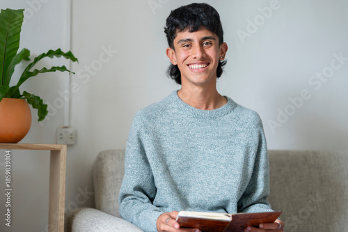 Young man smiling while reading a book sitting on a sofa at home