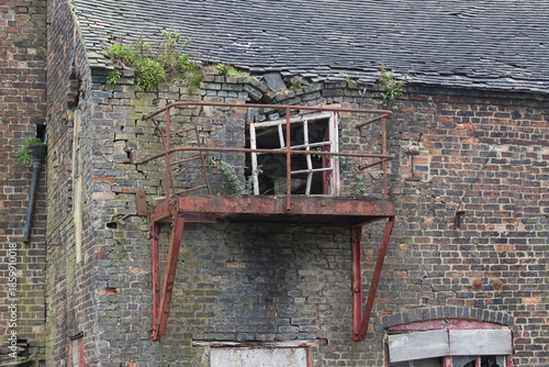 a close up of an old derelict window located in an old disused building.The bricks and roof are falling and the metal balcony is rusty. Vegetation is growing out of the brickwork