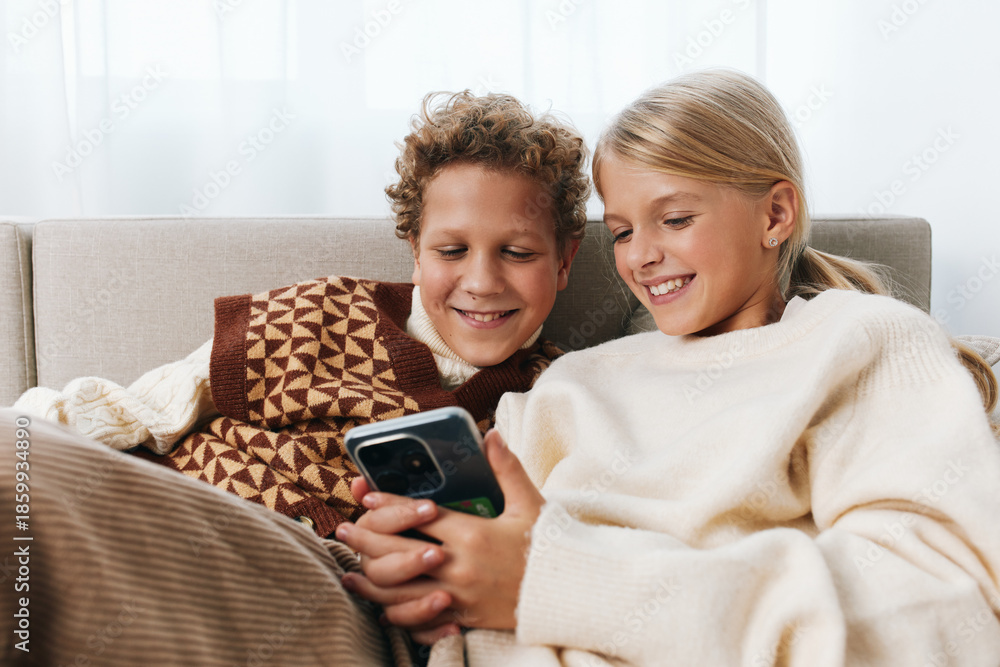 Fototapeta premium Authentic moment of playful learning and family lifestyle as two children share a cozy screen moment on the sofa, smiling together in a warm, relaxed setting.
